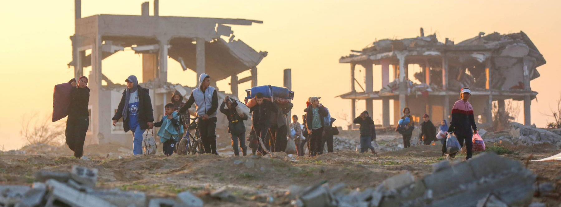 A group of people carrying their few belongings, walking in front of buildings in ruins, at sunset.